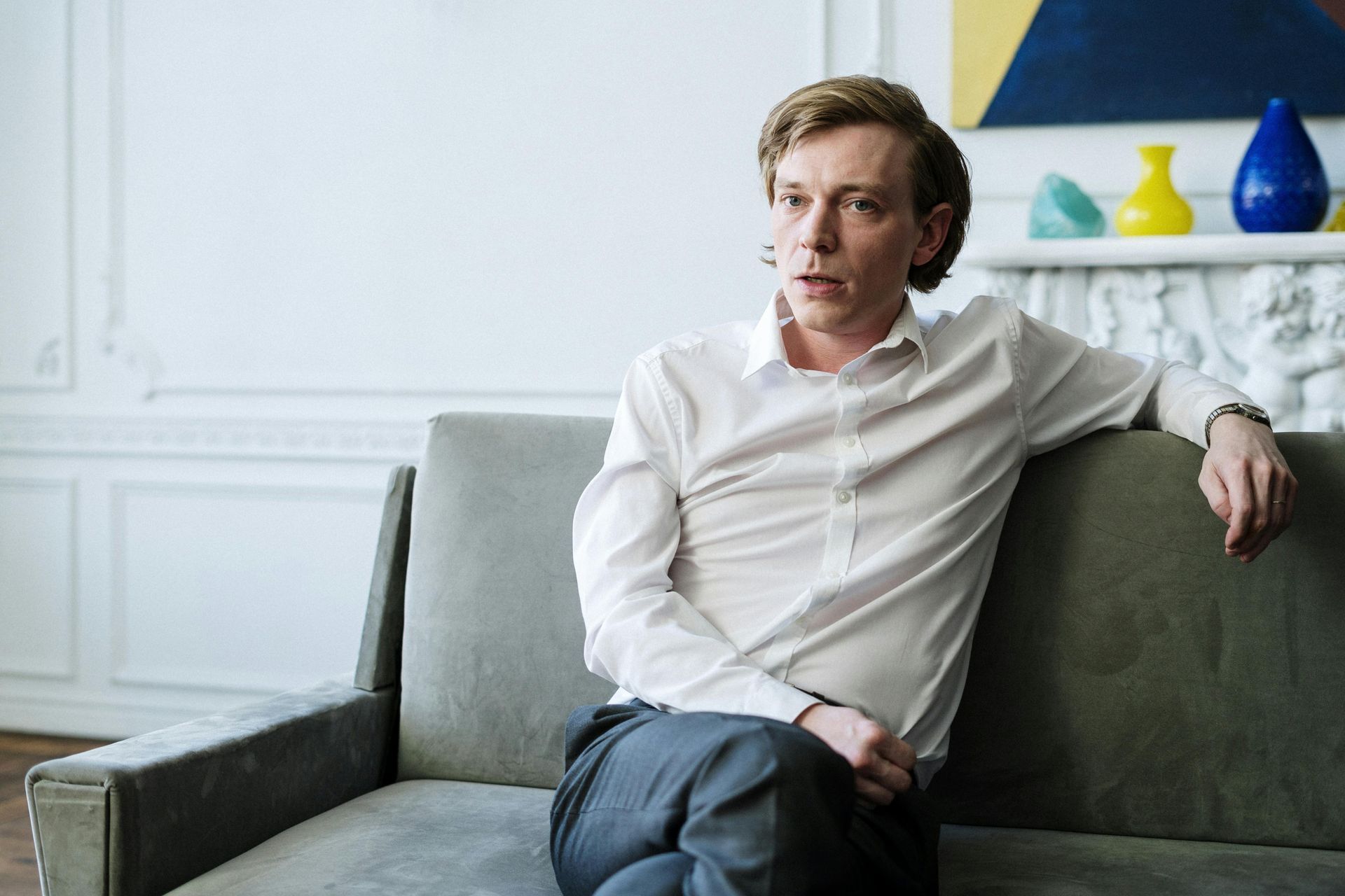 Man with light skin sitting on a gray couch, wearing a white shirt and gray pants, in a room with art.