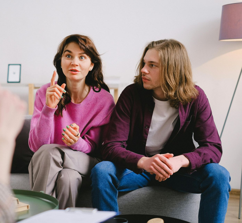Woman gesturing, speaking to a therapist while teen boy looks on, sitting in a counseling session.