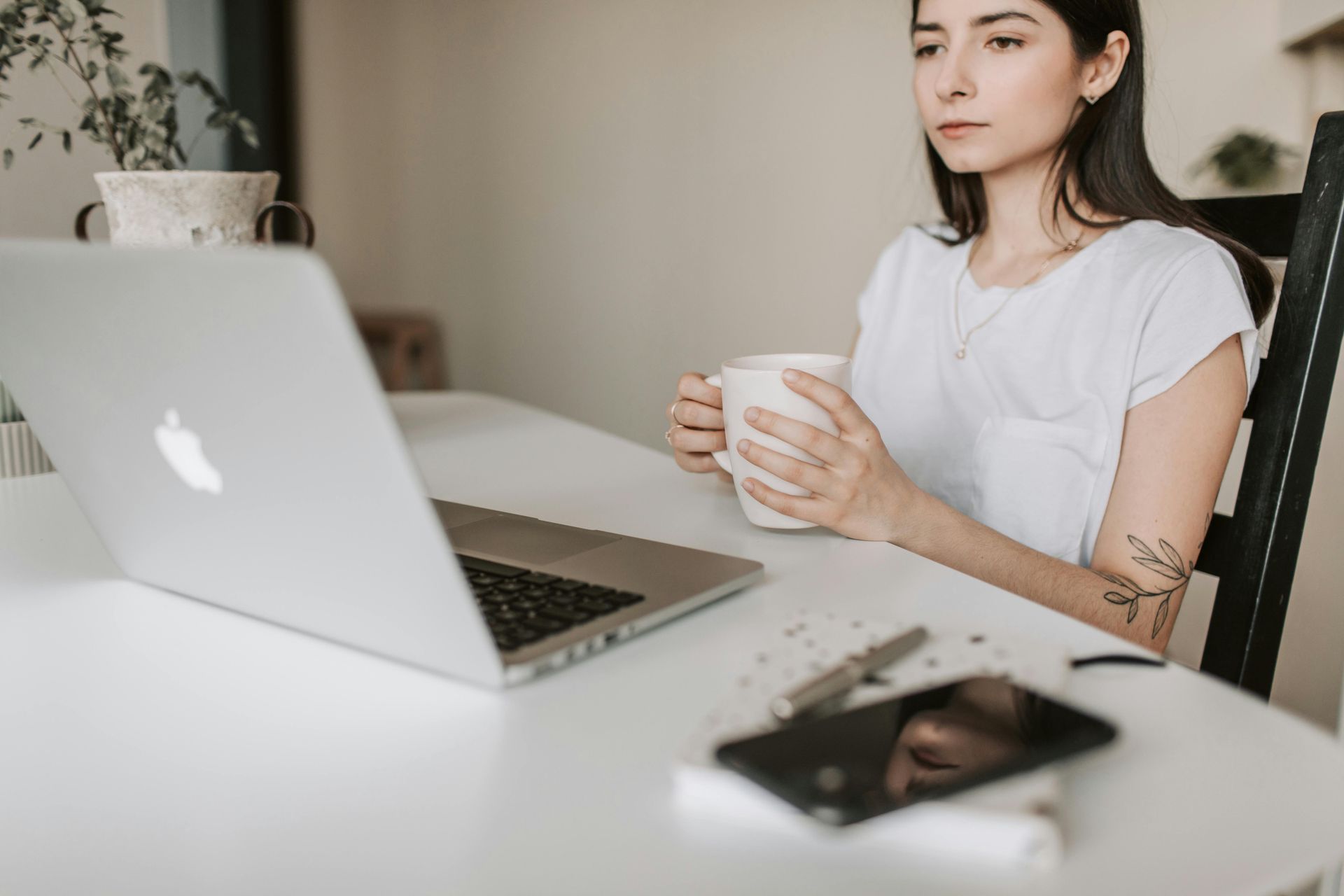 Woman with mug using laptop at table; pen and phone visible.