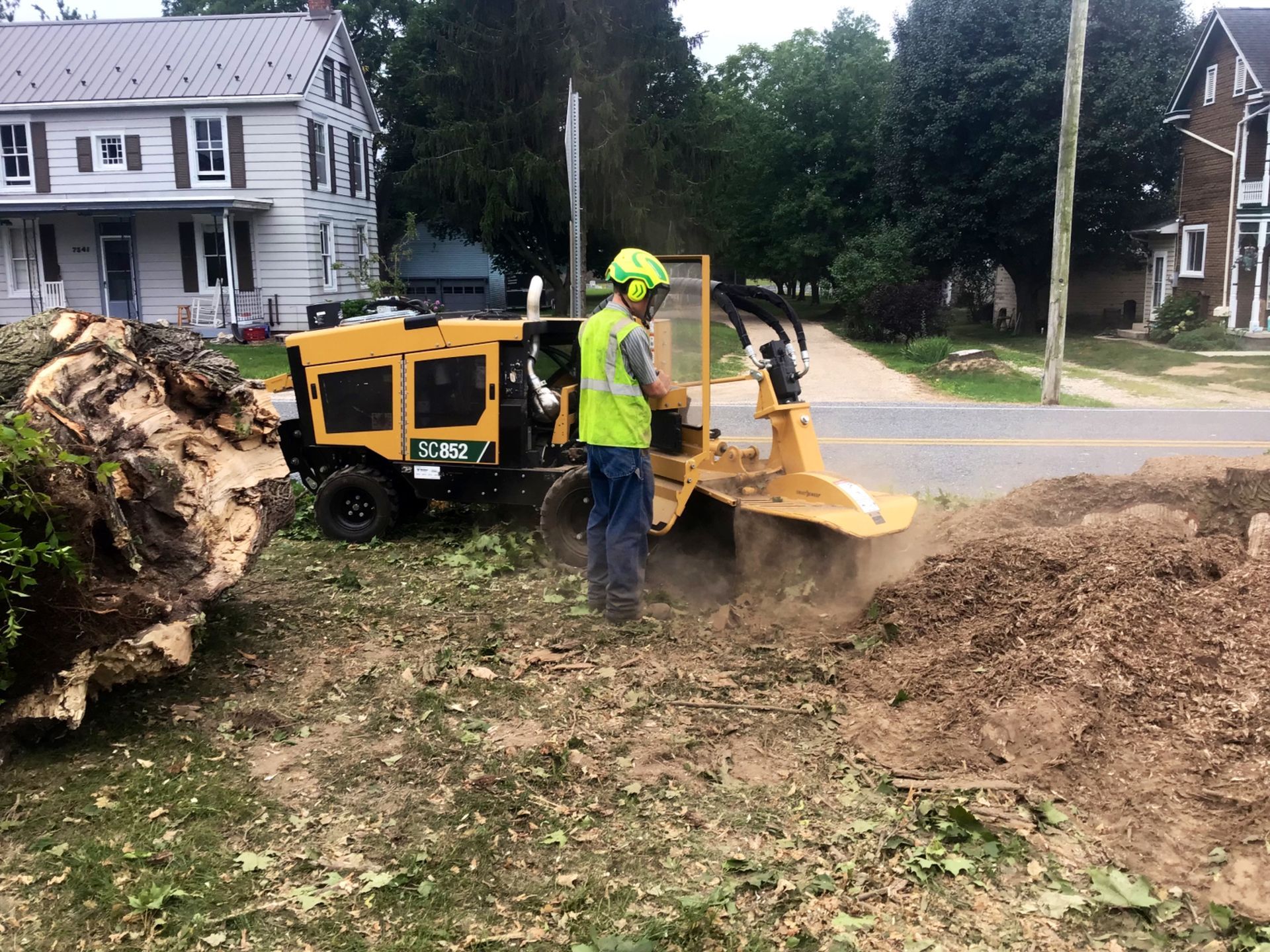 Man operating a stump grinder; grinding wood on residential lawn, next to a street.