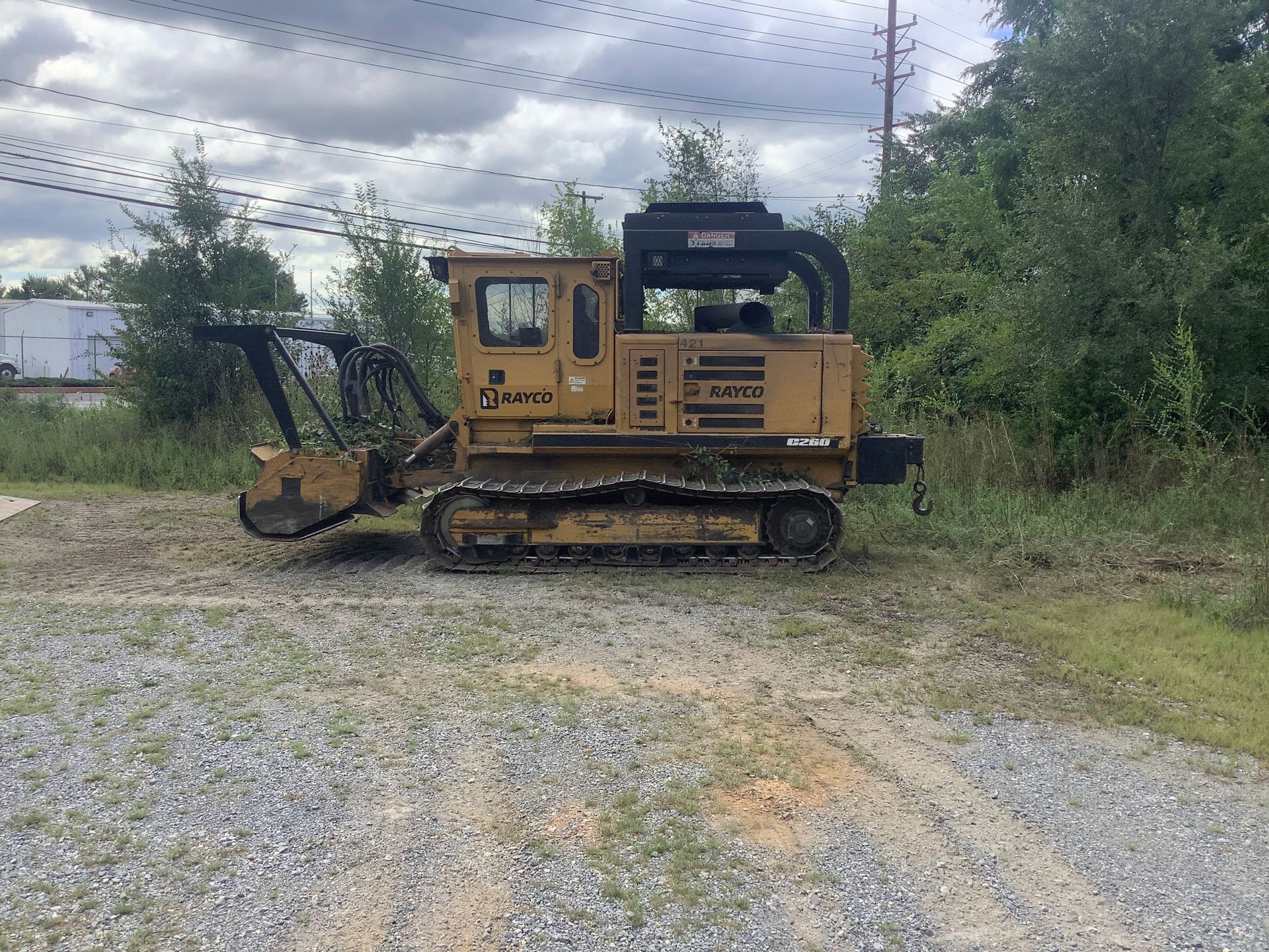Yellow forestry mulcher on a gravel area, with trees and power lines in the background.