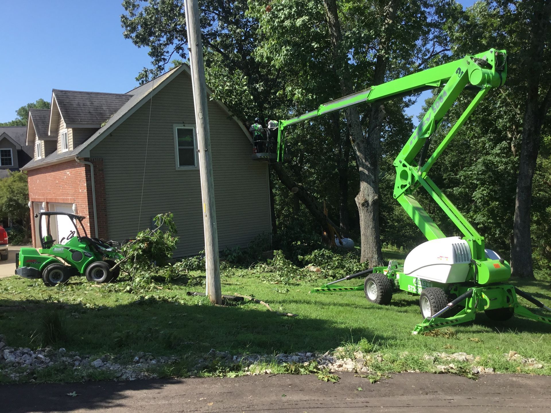 Green lift cutting tree branches near a house. Green chipper on the left.