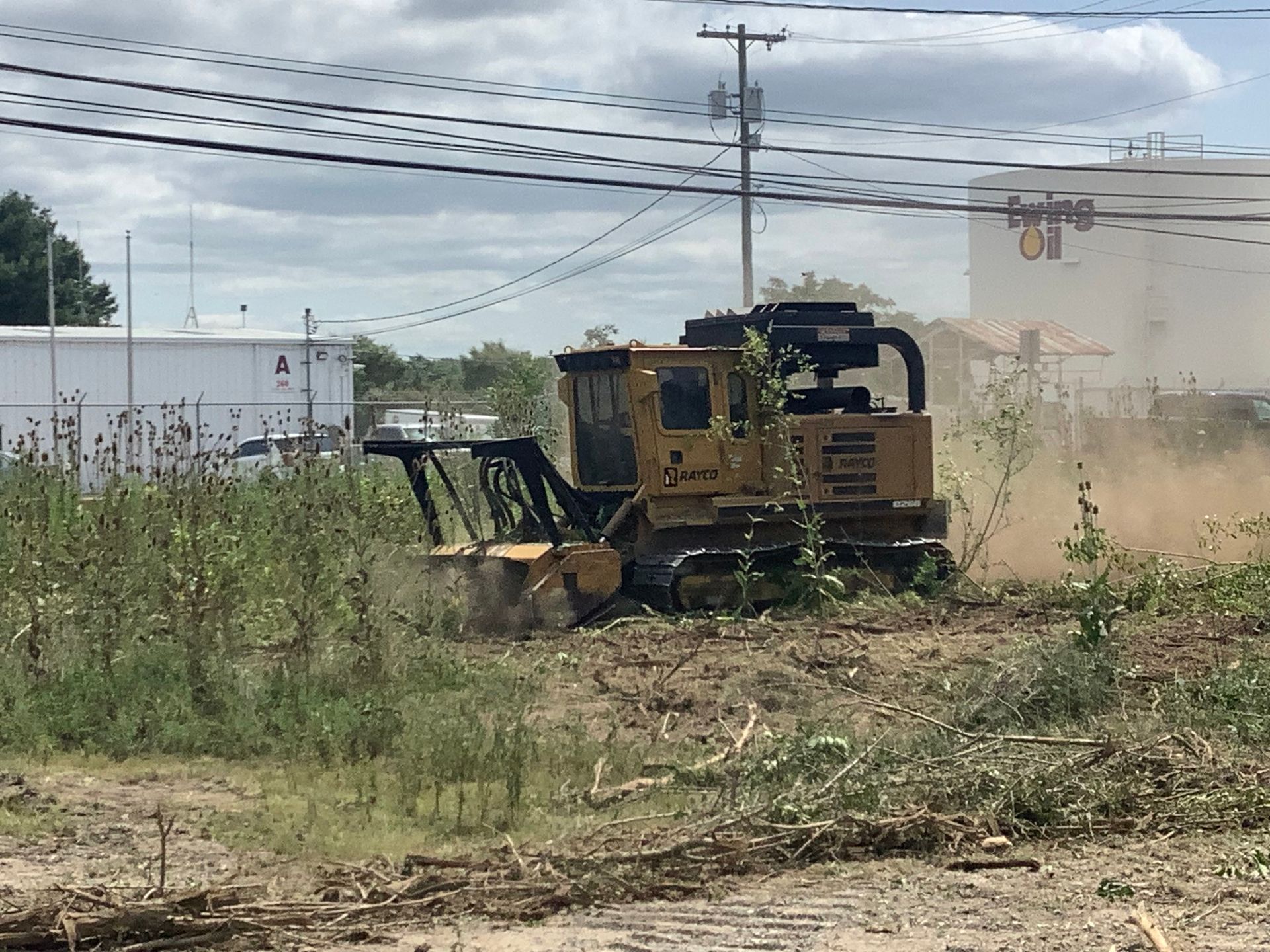 Yellow forestry mulcher clearing brush in an industrial area, dust clouds rising.