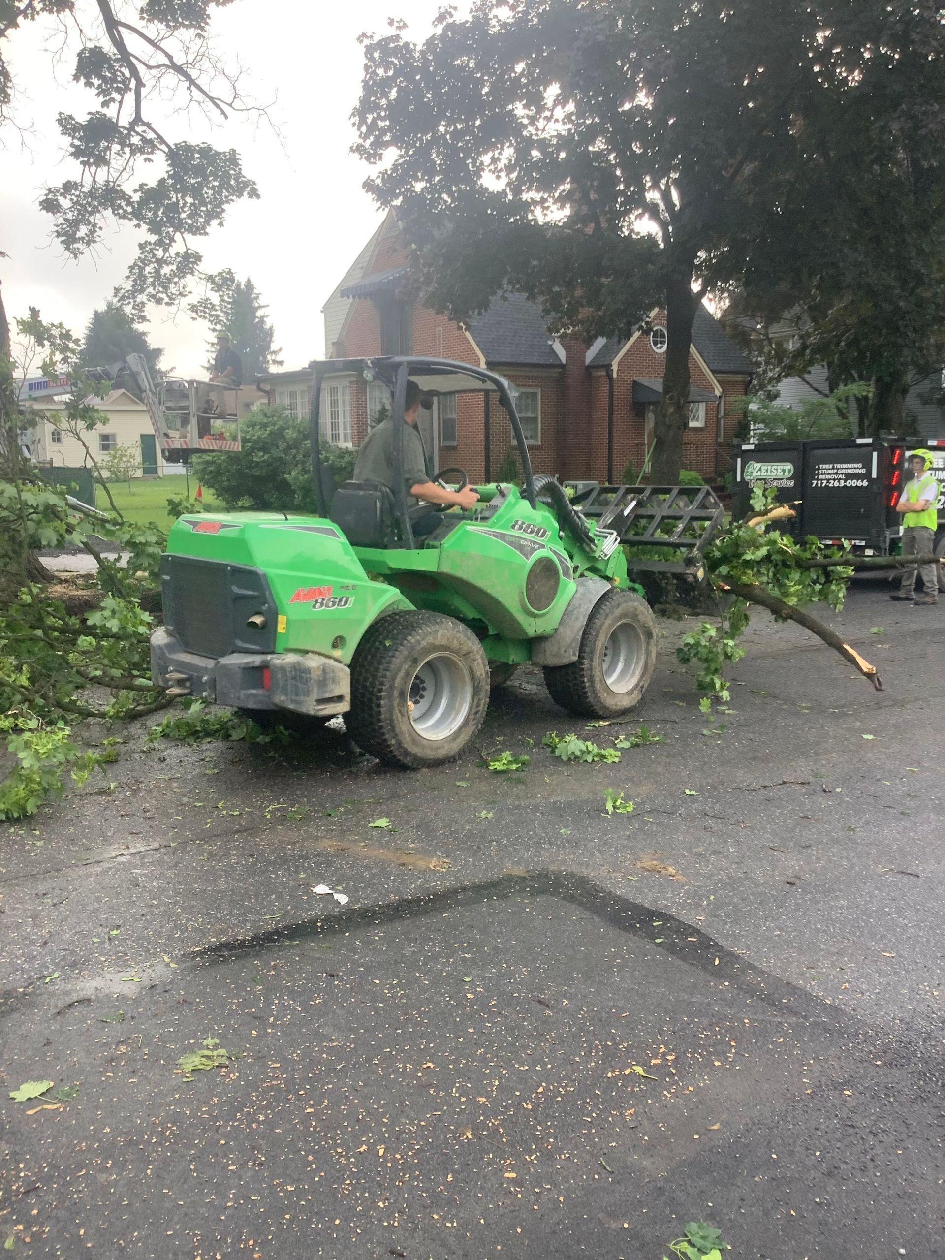 Green utility vehicle clearing tree branches from a street, with a worker in the cab.