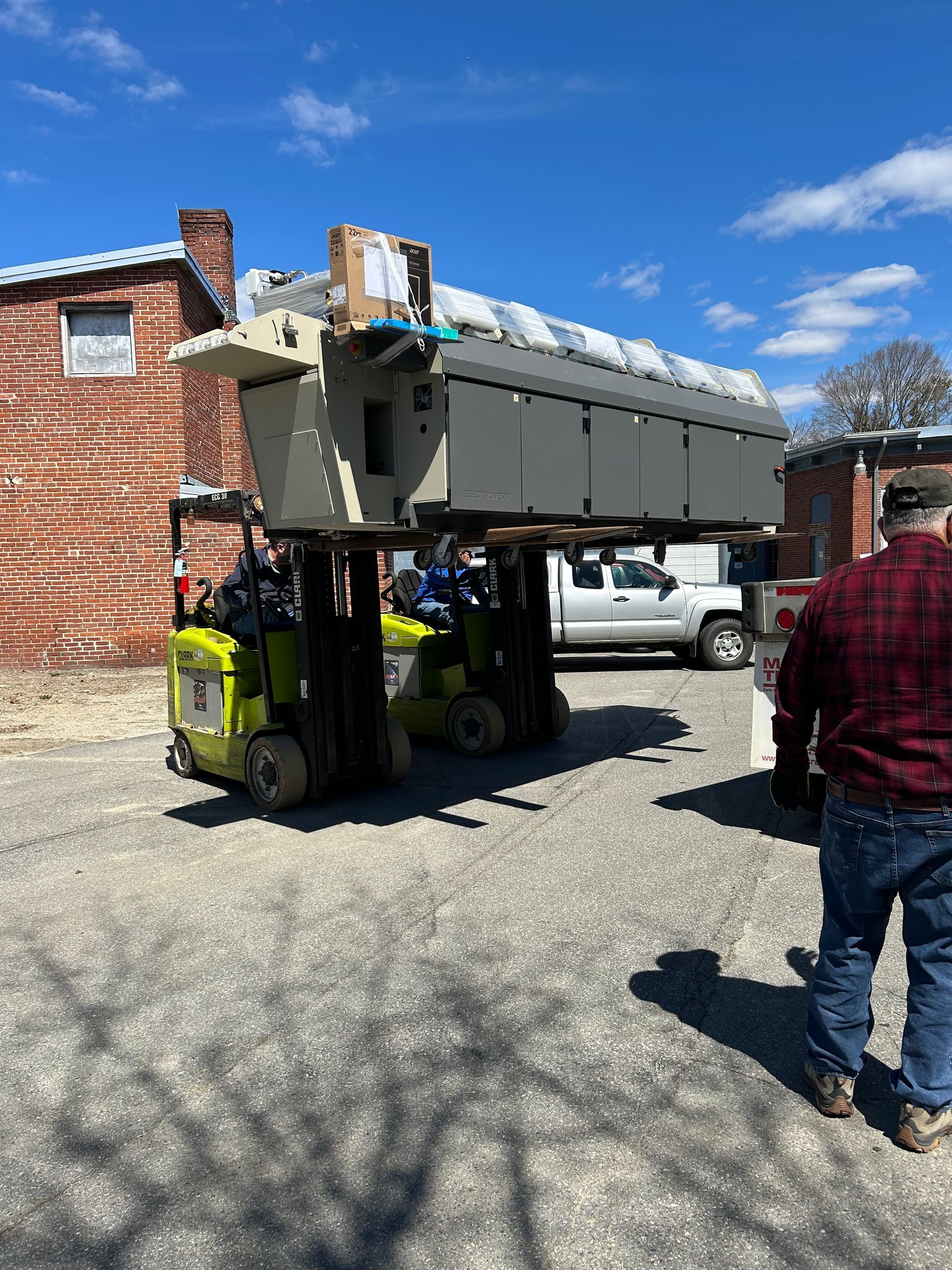 A man in a plaid shirt is standing next to a forklift carrying a large object.