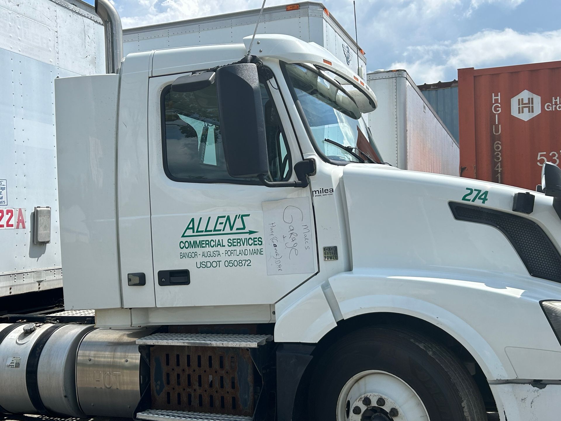A white semi truck is parked in front of a row of shipping containers.