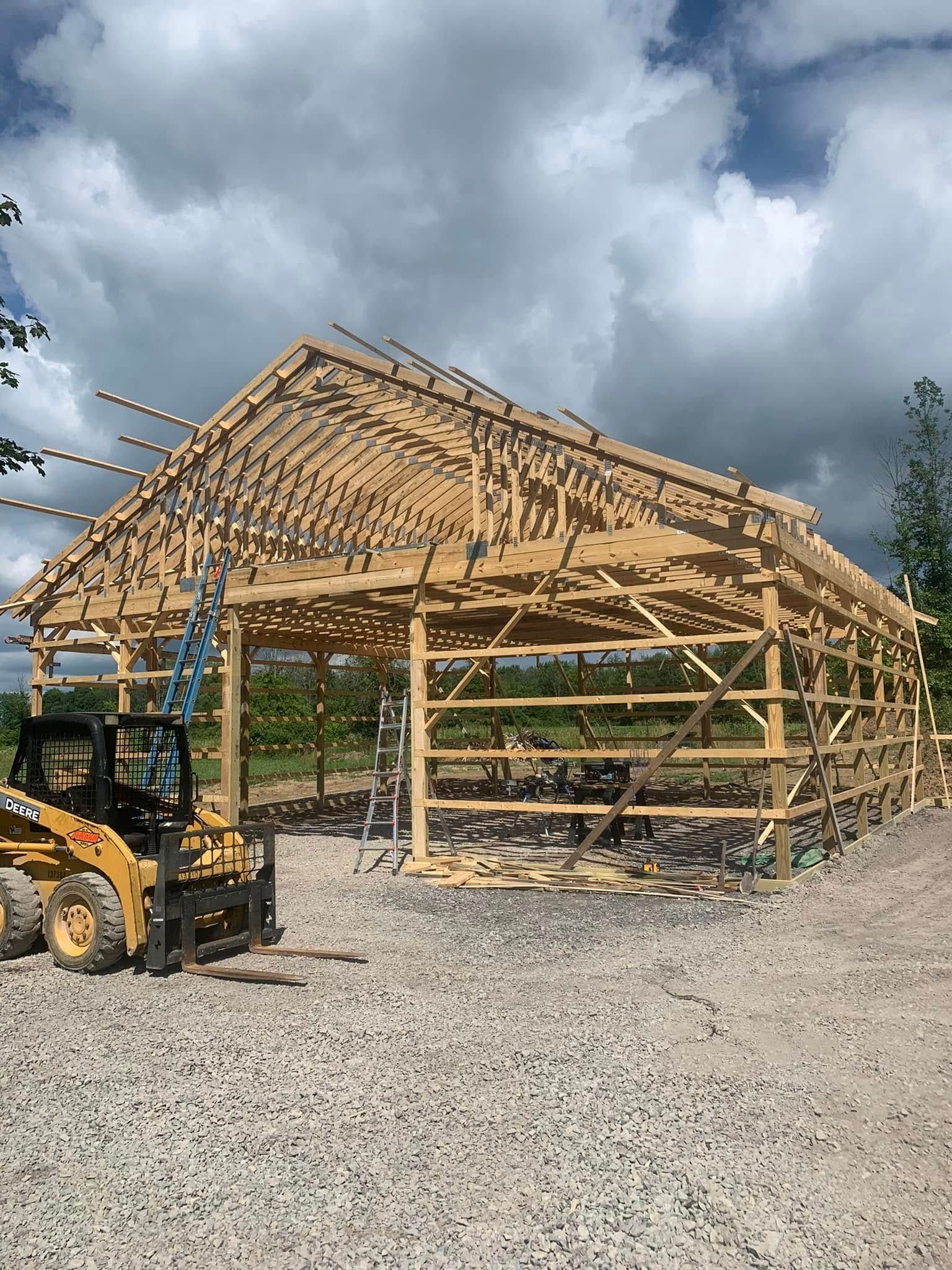 Construction of a wooden building frame, with a yellow front-end loader in the foreground, on a gravel lot under a cloudy sky.
