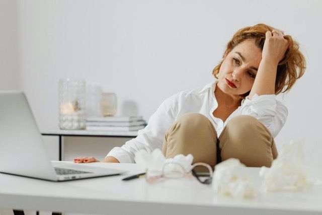 Woman looking stressed, sitting at a desk with a laptop, hand in hair. Beige pants, white shirt.