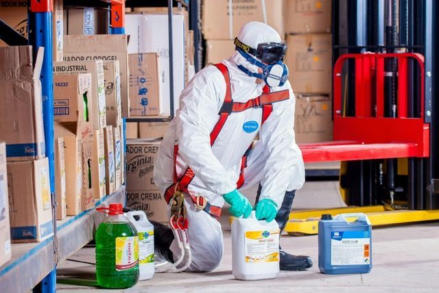 Person in hazmat suit kneels in a warehouse, handling chemical containers.