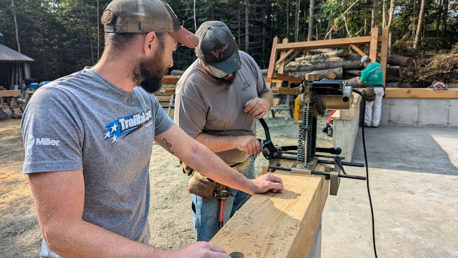 Two workers measuring lumber on a construction site with tools and framing in the background