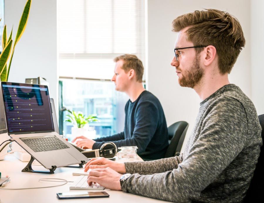 Two men working on laptops in an office, one wearing glasses and typing, the other in the background.