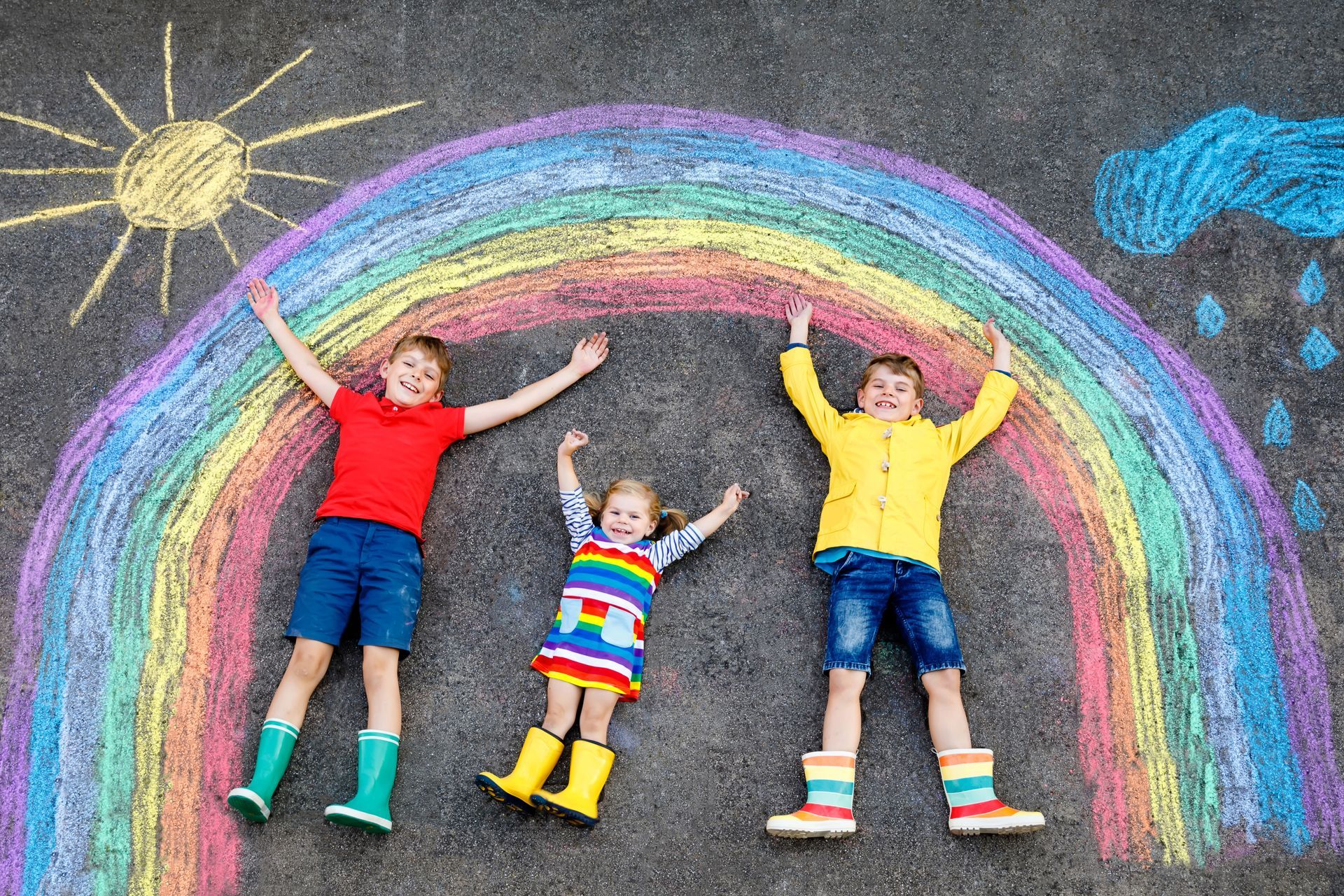 Children lying under a rainbow drawn on pavement, with sun and rain clouds.