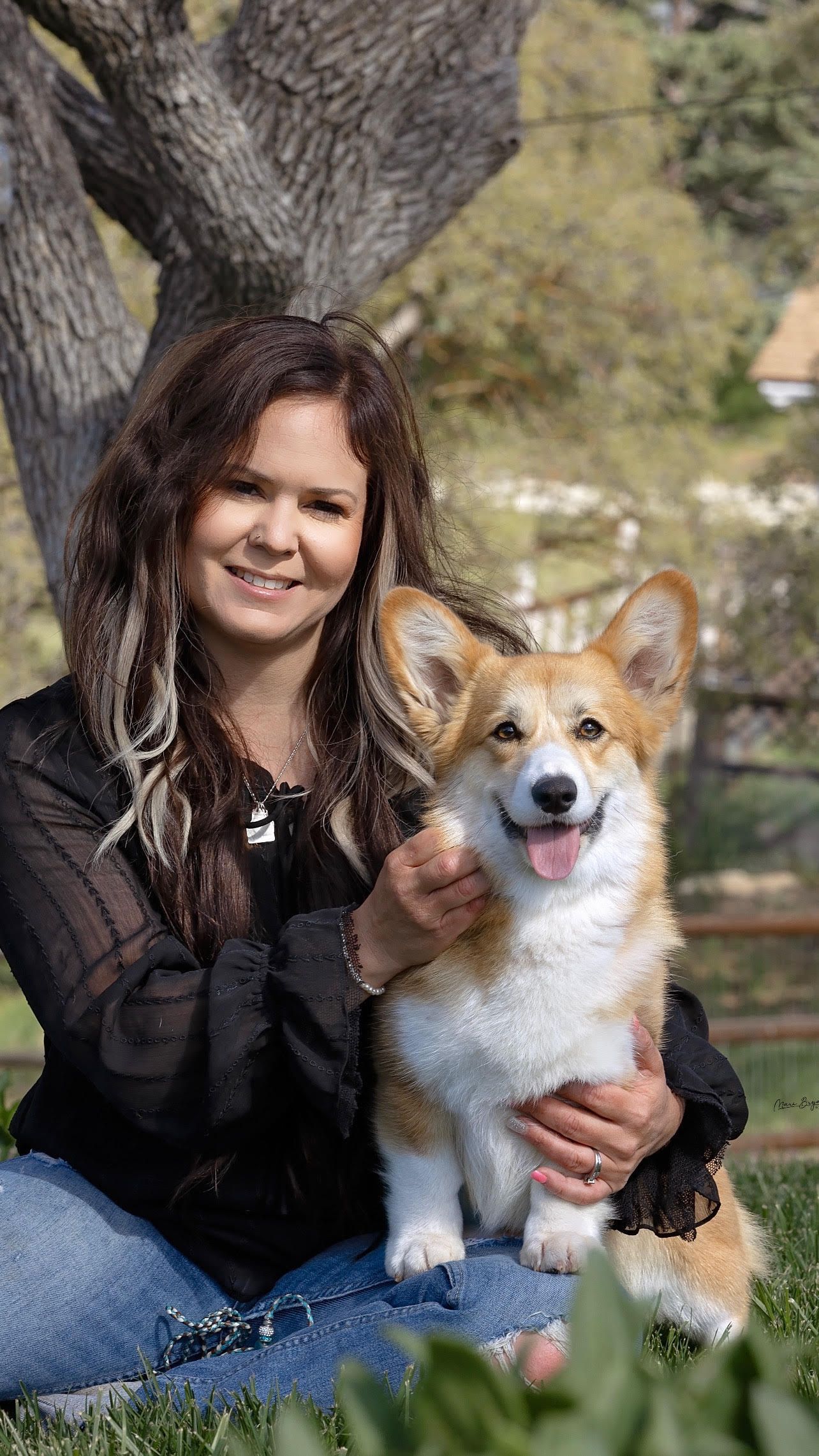 A woman is sitting on the grass holding a corgi dog.