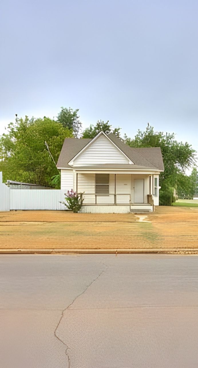 Small white house with porch, brown roof, in front of dry grass and a white fence.