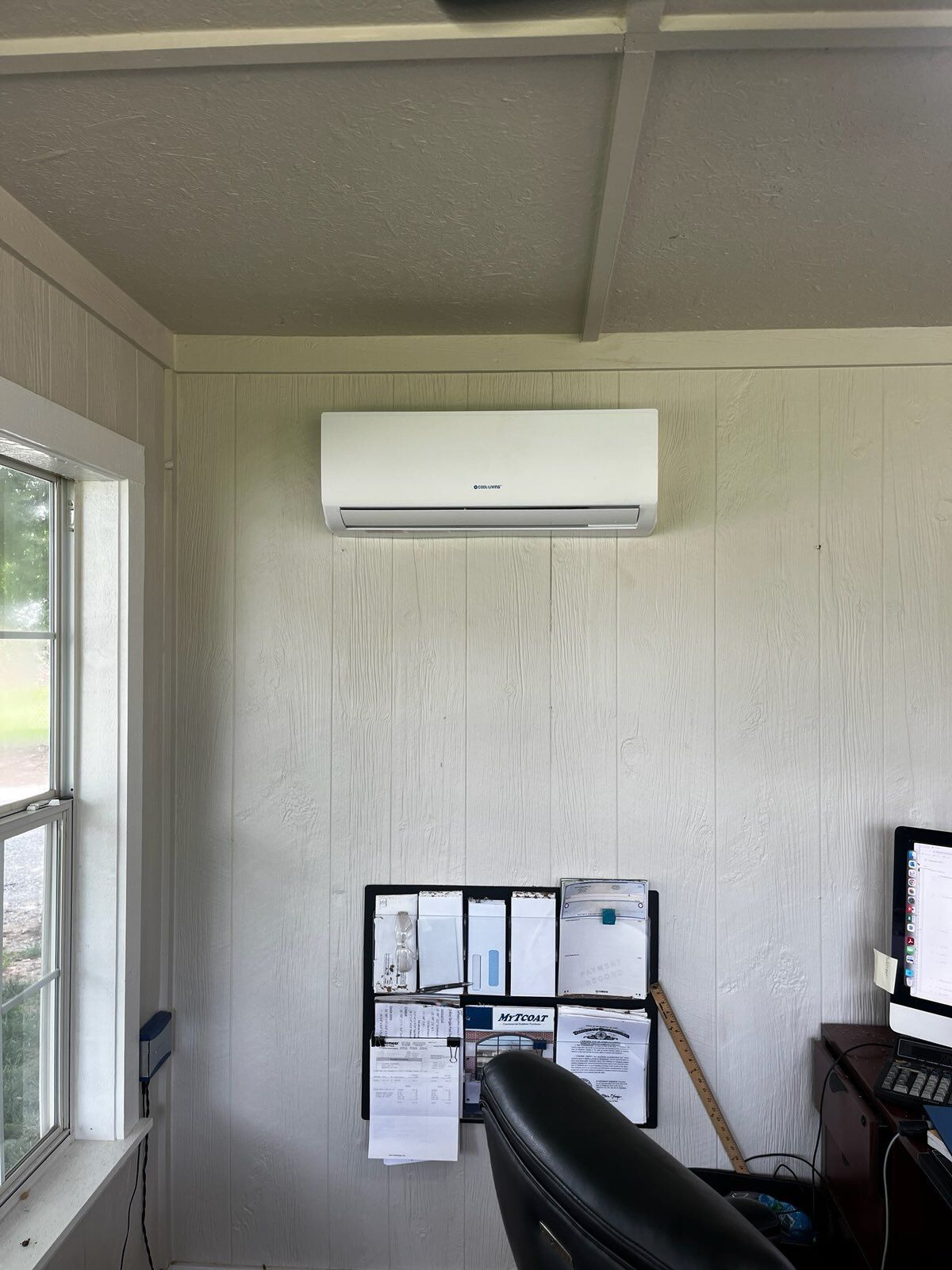 White air conditioning unit mounted on a white paneled wall above a desk in an office.