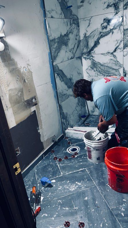 Person tiling a bathroom. White, gray, and blue marble-like tiles. Buckets on the floor.