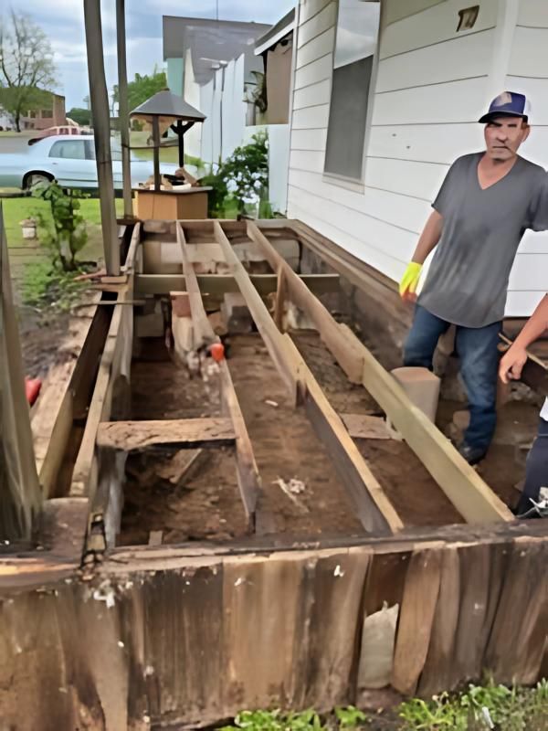 Men rebuilding a wooden porch with exposed framing. One man wears a hat and jeans. Outdoors.