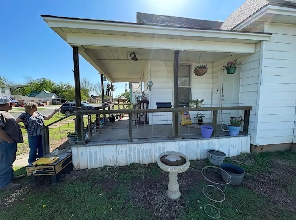 People on porch of white house with plants, bird bath, and yard. Blue sky.