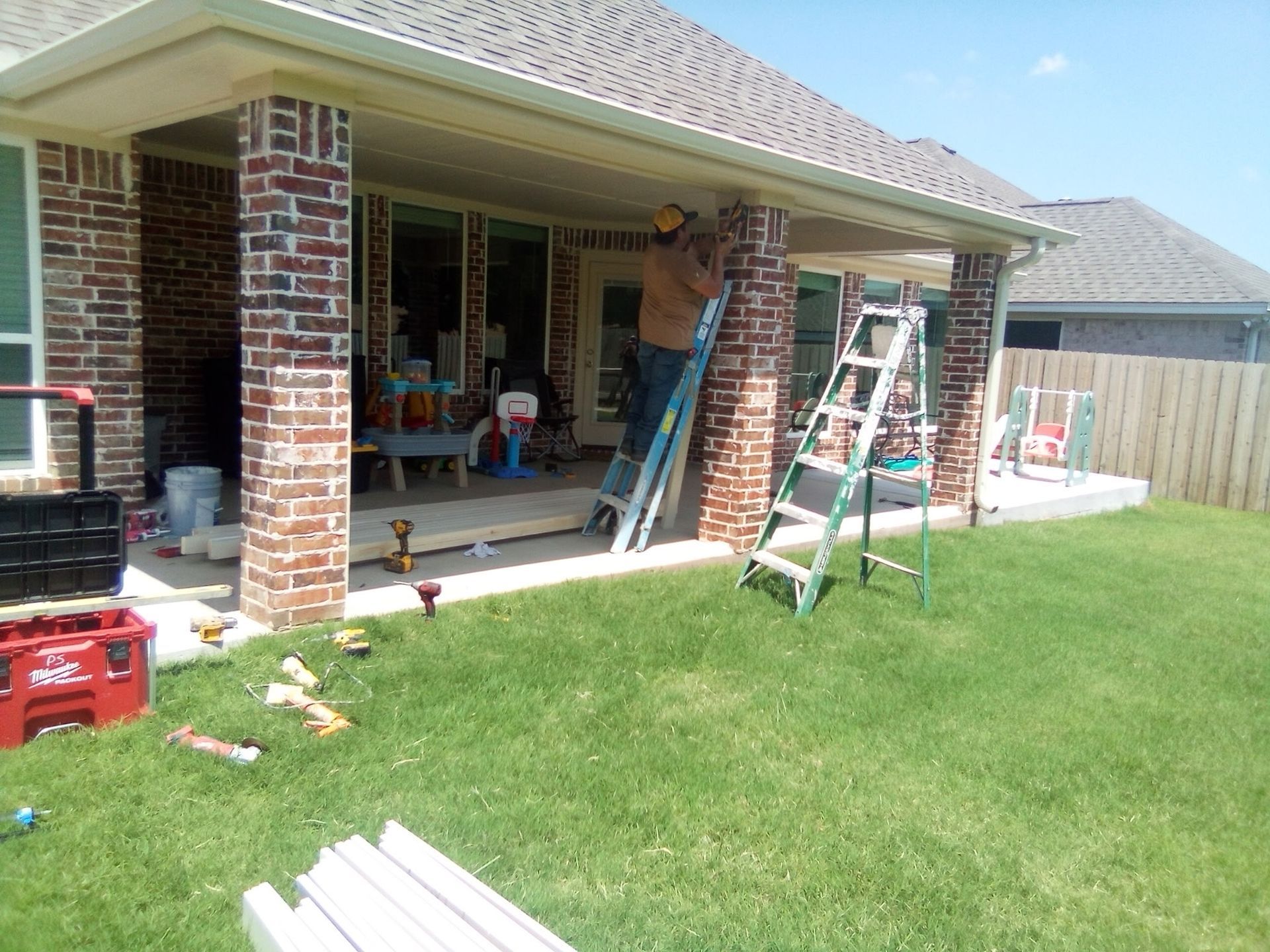 Man on ladder installing trim on a brick porch, with tools and materials scattered in the yard.