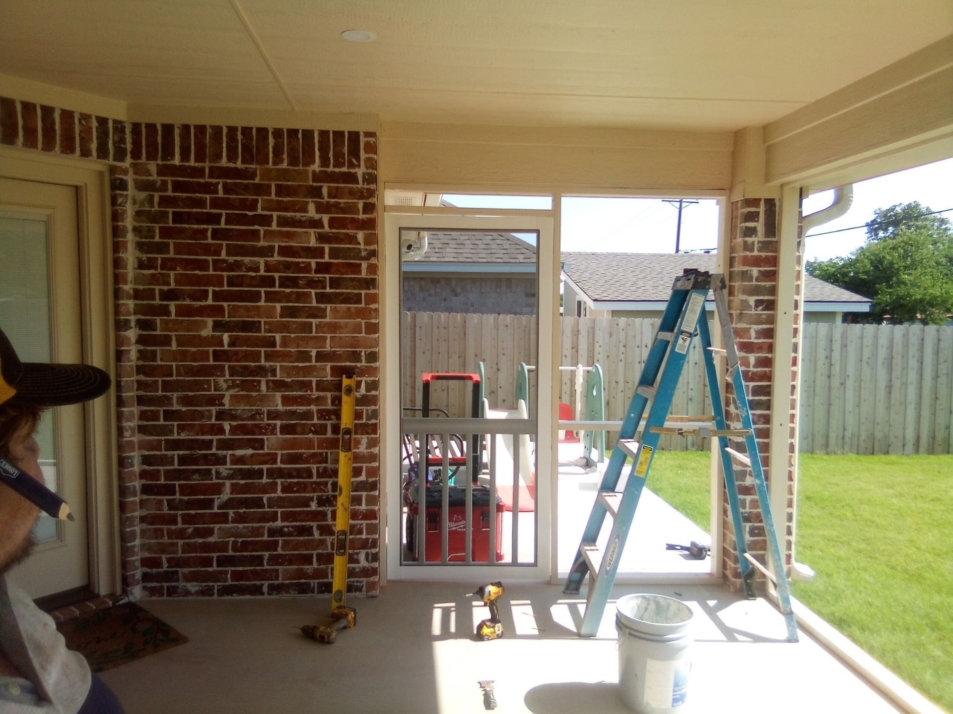 Screened-in porch with brick wall, a ladder, and a door. Bright sunlight illuminates the space.