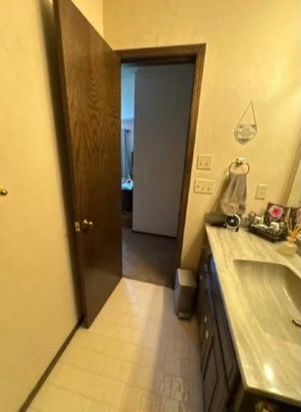 Bathroom with open door to hallway, vanity, brown door and cabinets, beige walls and floor, towel rack.