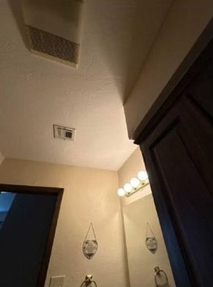 Bathroom interior: vent, light fixtures, mirror, door, and hanging decor on cream-colored walls.
