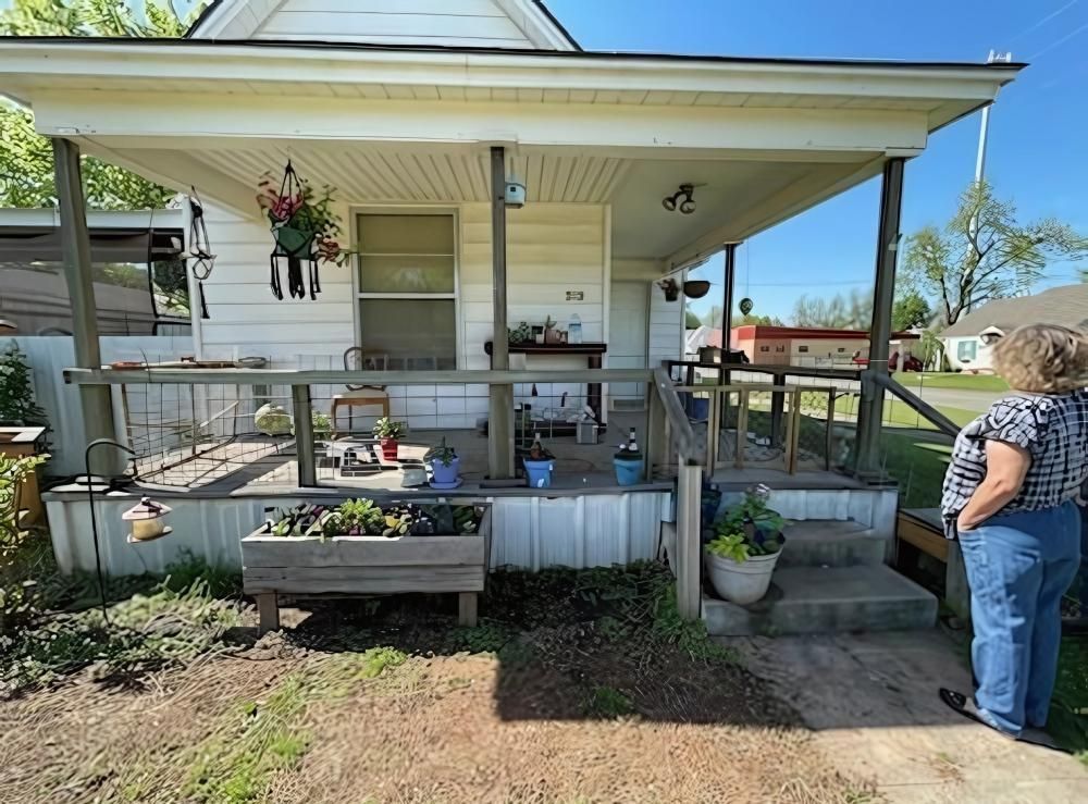 White house with porch, railing, woman looking on the right. Plants, pots, and hanging decor.
