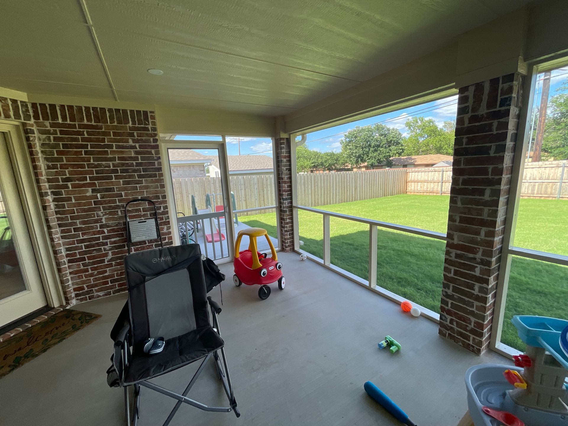 Screened-in porch with a brick wall, overlooking a green backyard; has a child's car, chair, toys.