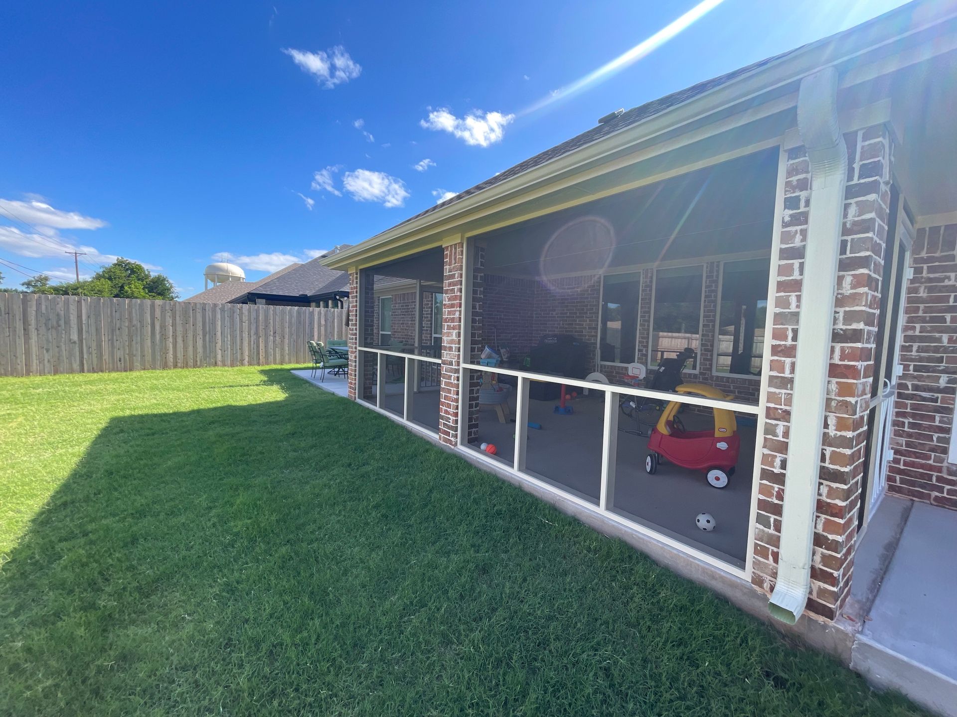 Screened-in porch with red brick columns, overlooking a green lawn and wooden fence on a sunny day.