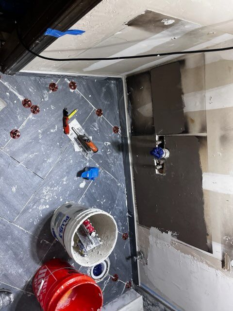 Bathroom under construction, gray herringbone tile on floor and partially on wall, plumbing and tools visible.