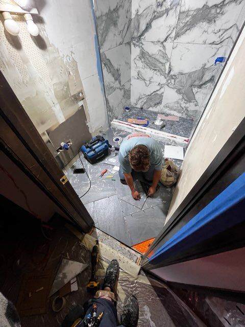 A person tiling a bathroom floor, viewed from a doorway. White and gray marble tiles are on the wall.