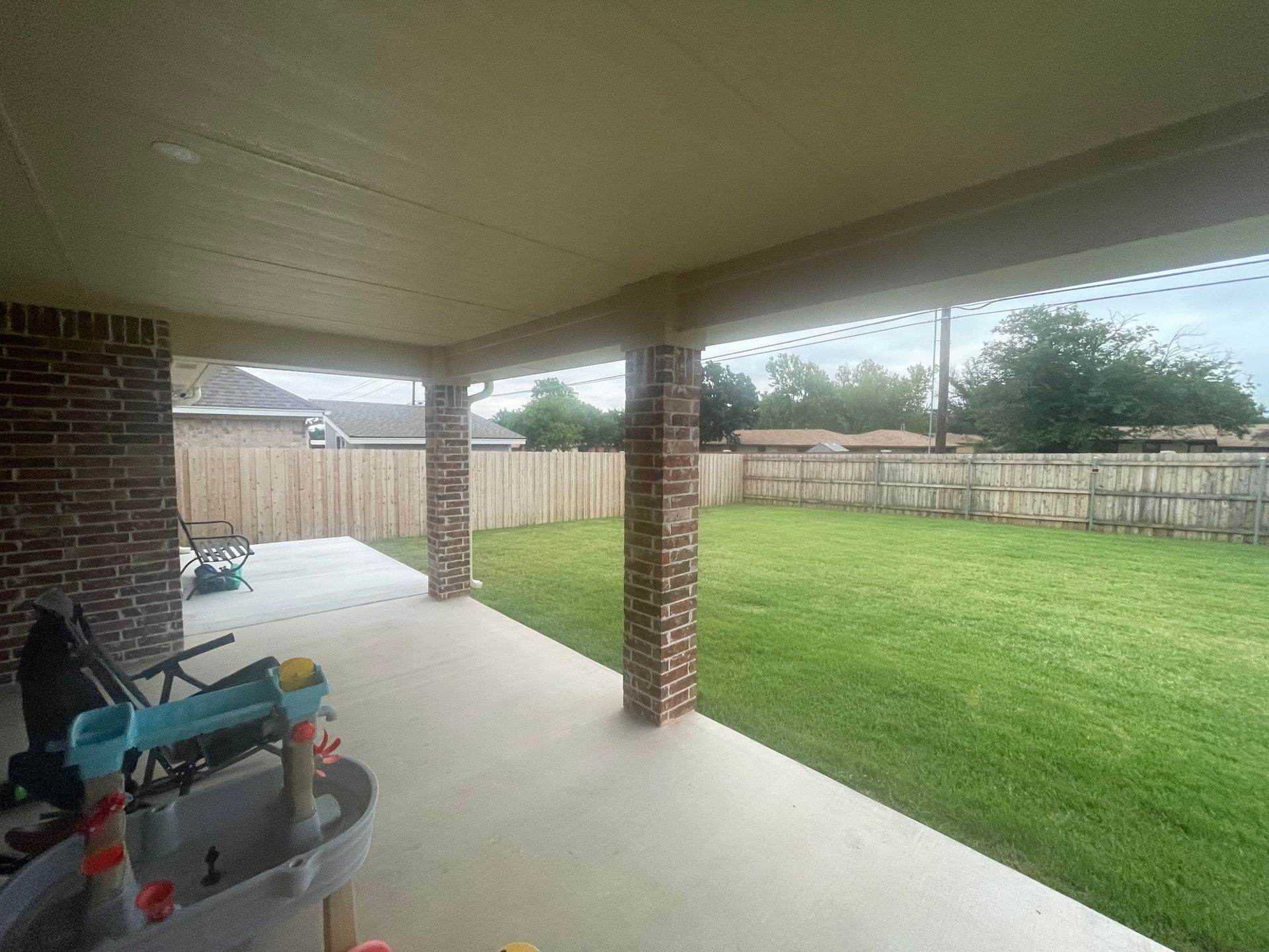 Covered patio with concrete floor, brick columns, and green lawn fenced in the background.