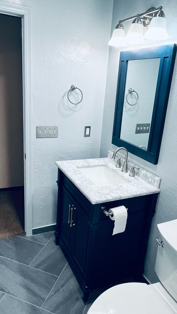Blue bathroom vanity with marble top, mirror, and toilet against a gray tiled floor and textured walls.
