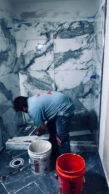 Man tiling a shower with marble-patterned tiles. Buckets and tools are on the floor.