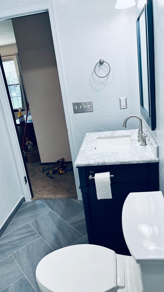 Bathroom with blue vanity, white toilet, gray tile, silver fixtures, and open doorway.