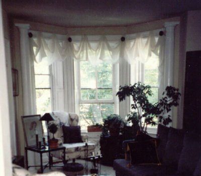 Bay window with draped white curtains, plants, and seating in a room.