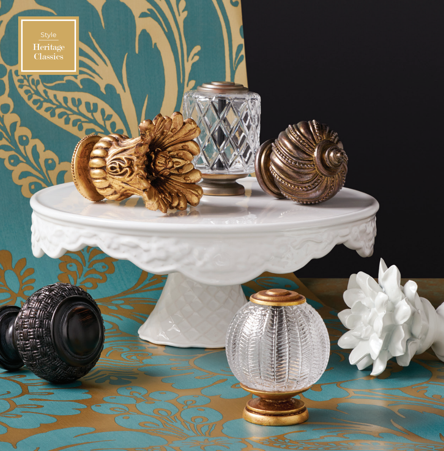 Various decorative knobs on a white cake stand, with teal and gold patterned wallpaper.