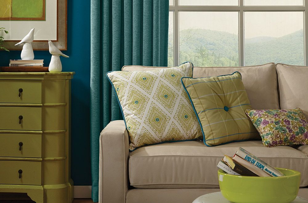 Beige couch with green and patterned pillows against teal wall, by window, next to green dresser.