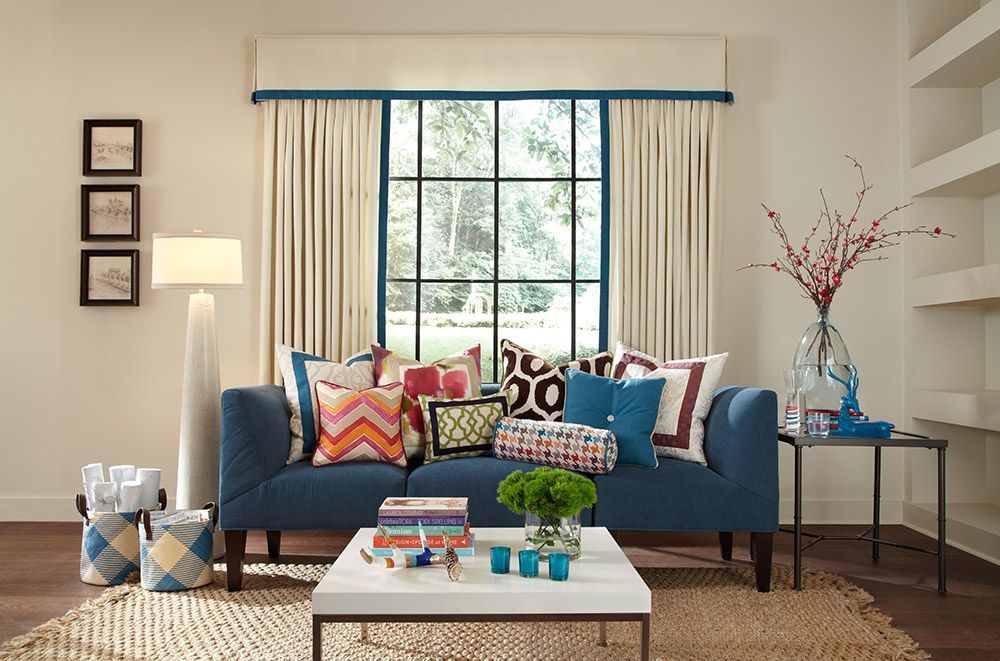 Blue sofa with colorful pillows in a living room; a window with curtains, table, and shelves.
