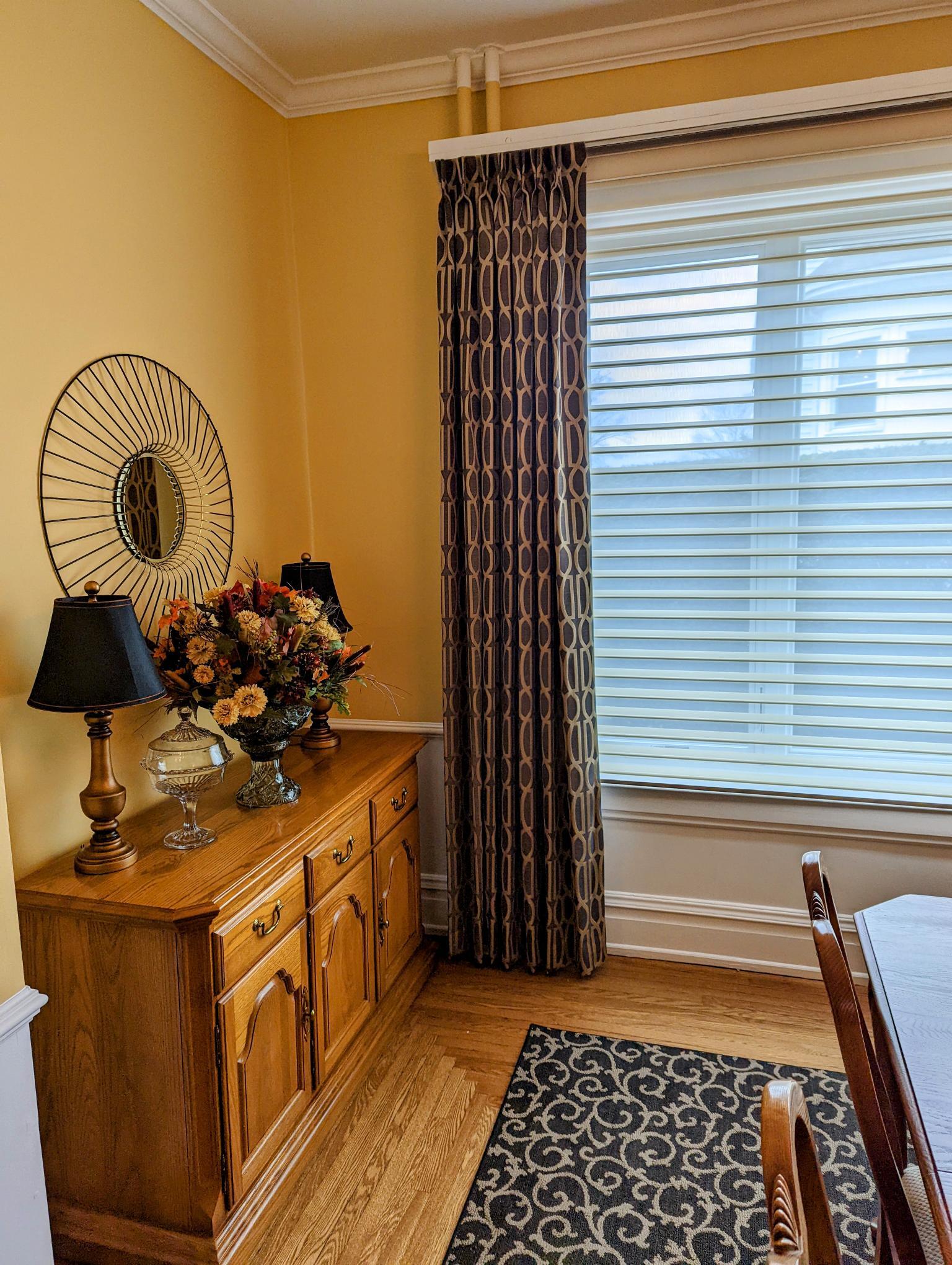 A dining room with yellow walls, wooden furniture, and a patterned rug near a window with blinds.