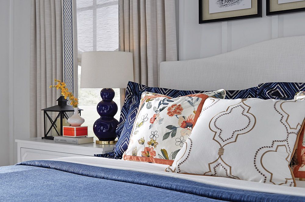 Bedroom with white headboard, blue bedding, decorative pillows, lamp, and bedside table.