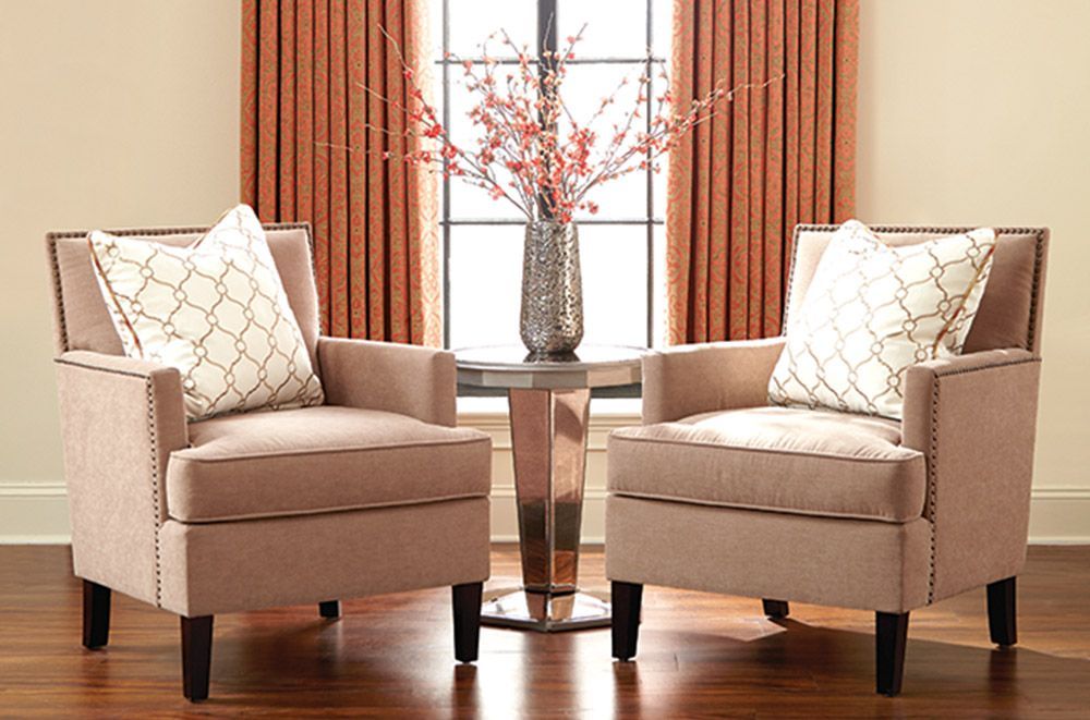 Two beige armchairs with decorative pillows facing a window with a vase, on a hardwood floor.