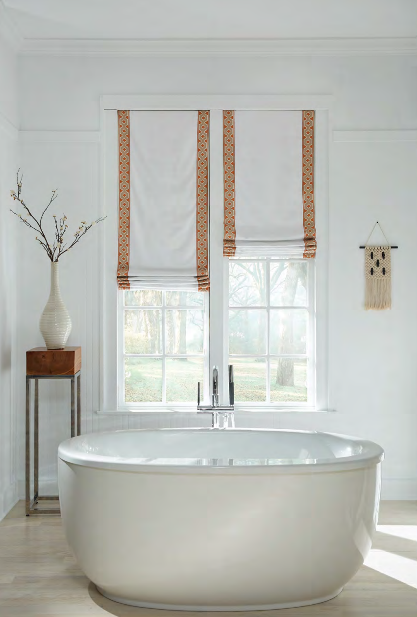 White bathroom with a freestanding tub, window with white shades, and a vase of flowers.