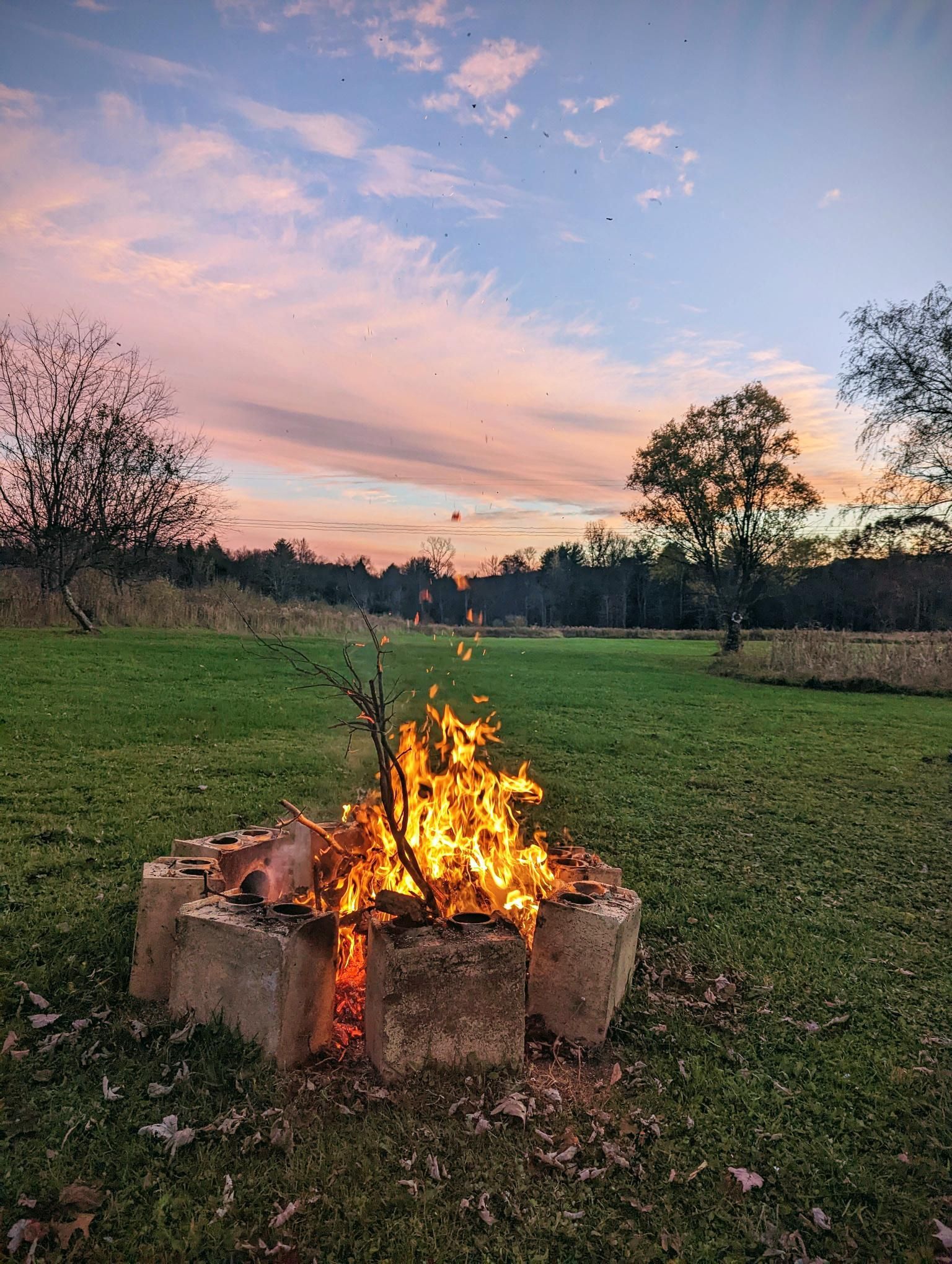 A campfire burns inside a concrete ring on a grassy field, under a pastel sunset.