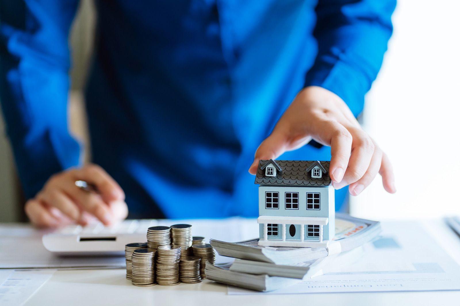 Person holding miniature house near coins and calculator, suggesting real estate planning.