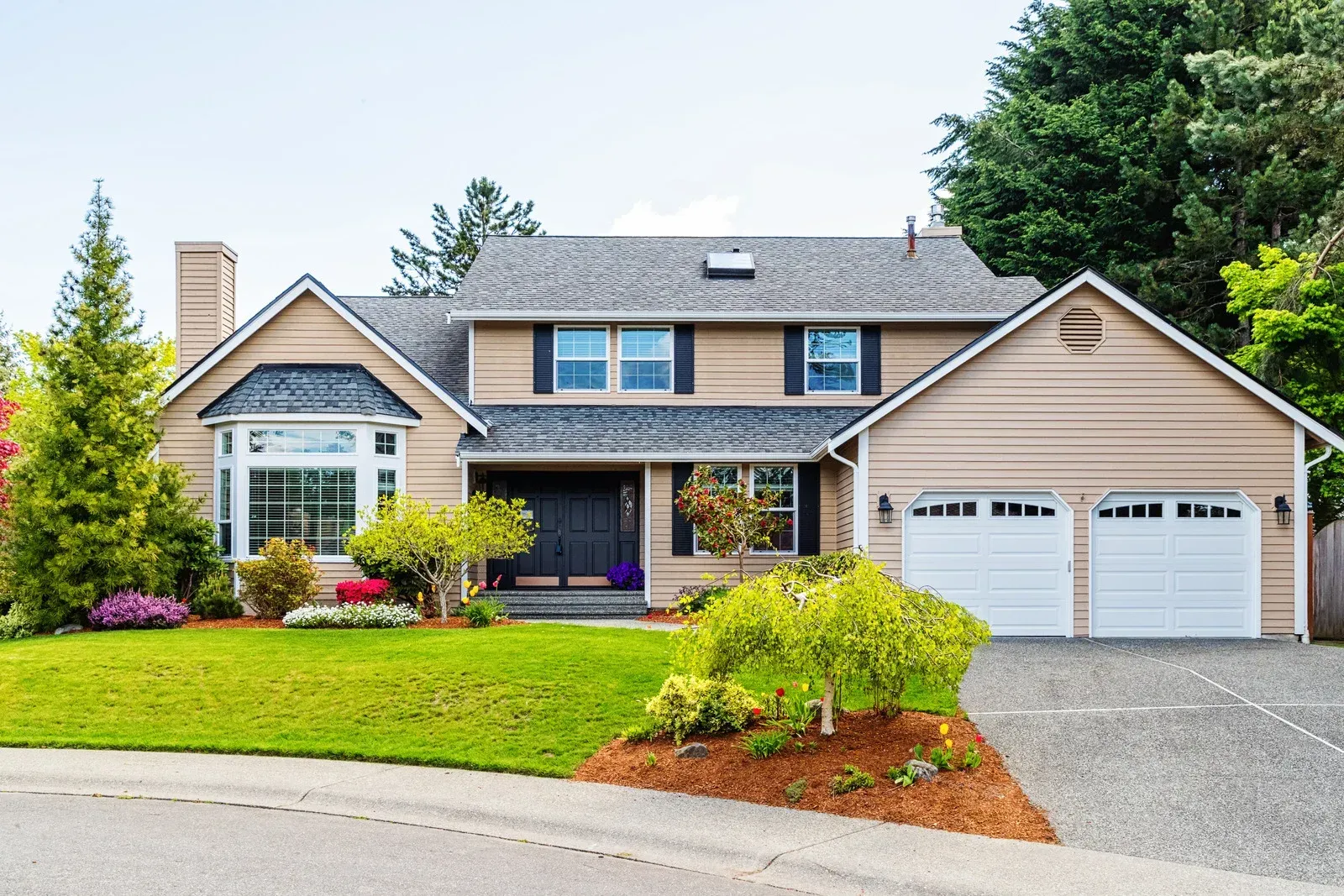 Two-story beige house with a green lawn and driveway. It has a bay window and a two-car garage.