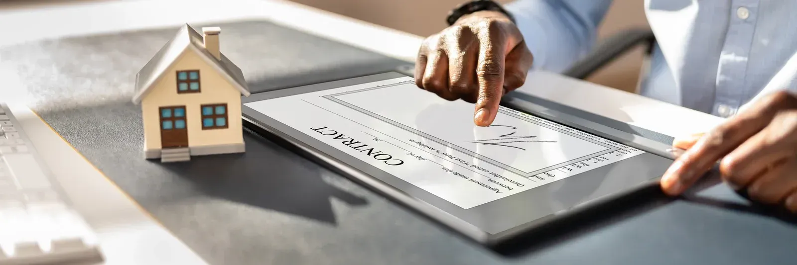 Person using a tablet to view or sign a check with a model house on the desk.