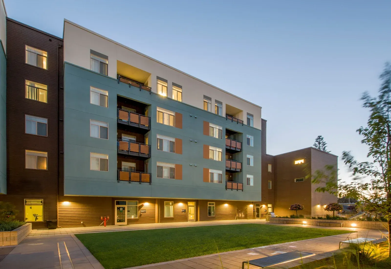 Modern apartment building with teal and brown siding, green lawn courtyard, and lit windows.