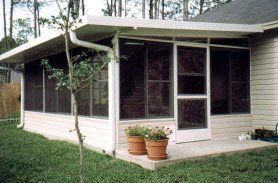 Screened-in porch attached to a light-colored house; beige siding, potted flowers, green grass.