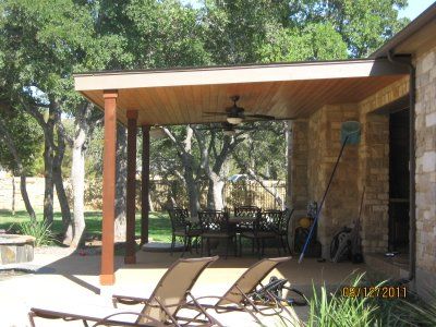 Outdoor patio with brown columns, a wooden ceiling, and stone wall, overlooking a yard with trees and furniture.
